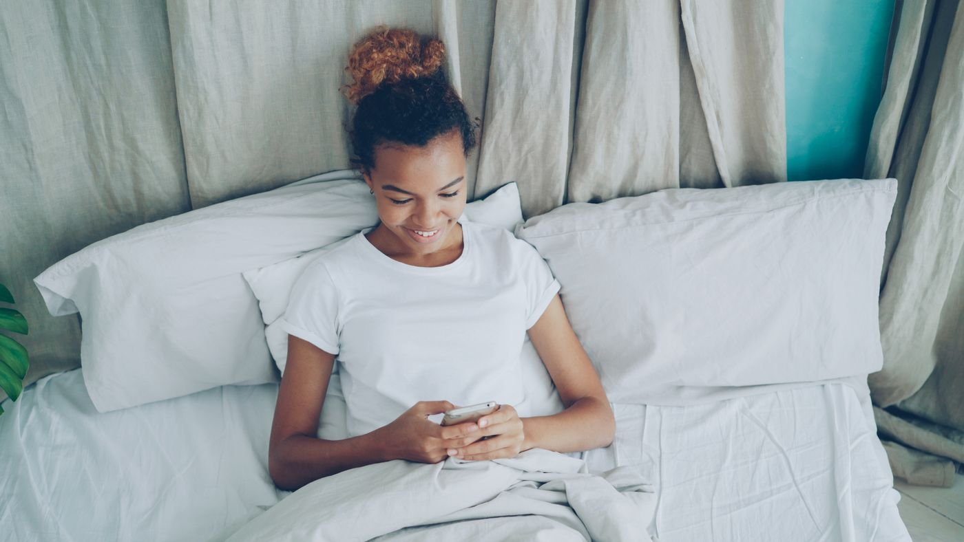 Woman smiling at her phone while texting her long distance partner from bed