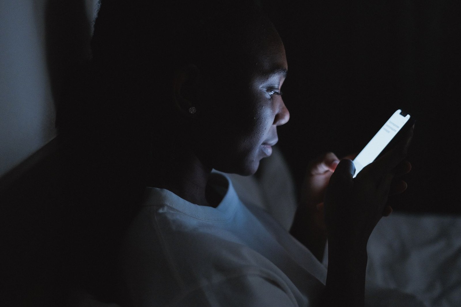 A woman checking a smartphone in a dark bedroom lit only by the screen