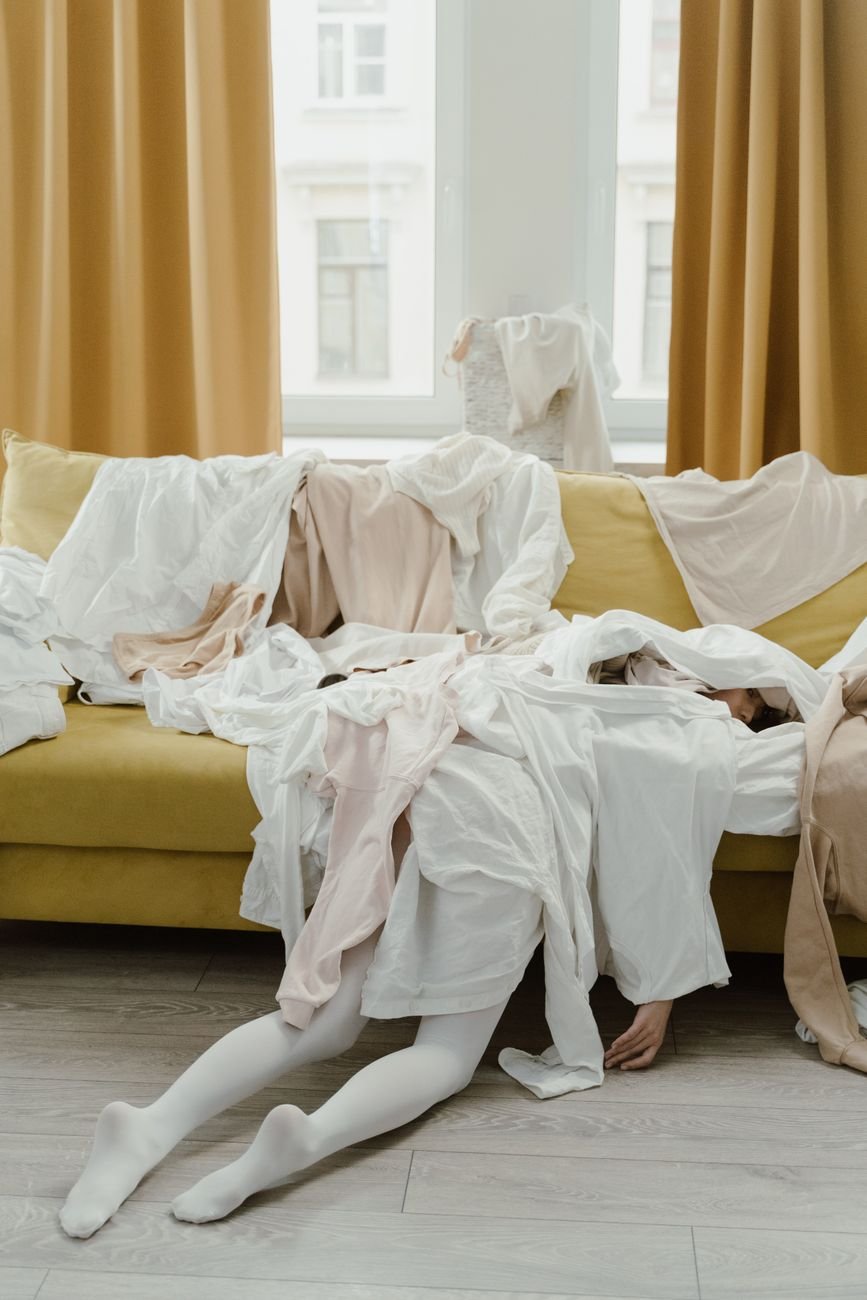 Woman sitting under a pile of unfolded clothes on a couch in a messy apartment